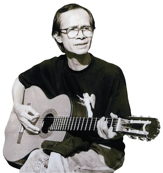 Black-and-white photo of Trịnh Công Sơn playing an acoustic guitar, wearing glasses and a black T-shirt