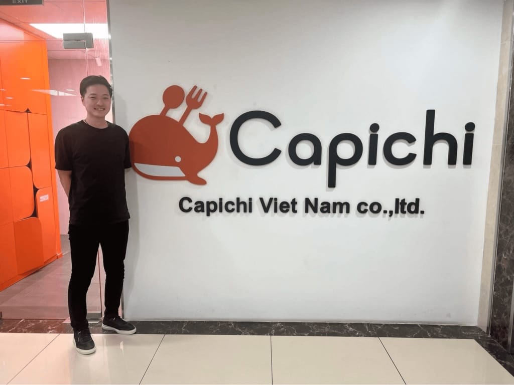 Capichi founder Mori Taiki standing next to the company's brand wall in the Hanoi office, with the orange whale logo and Capichi Viet Nam lettering