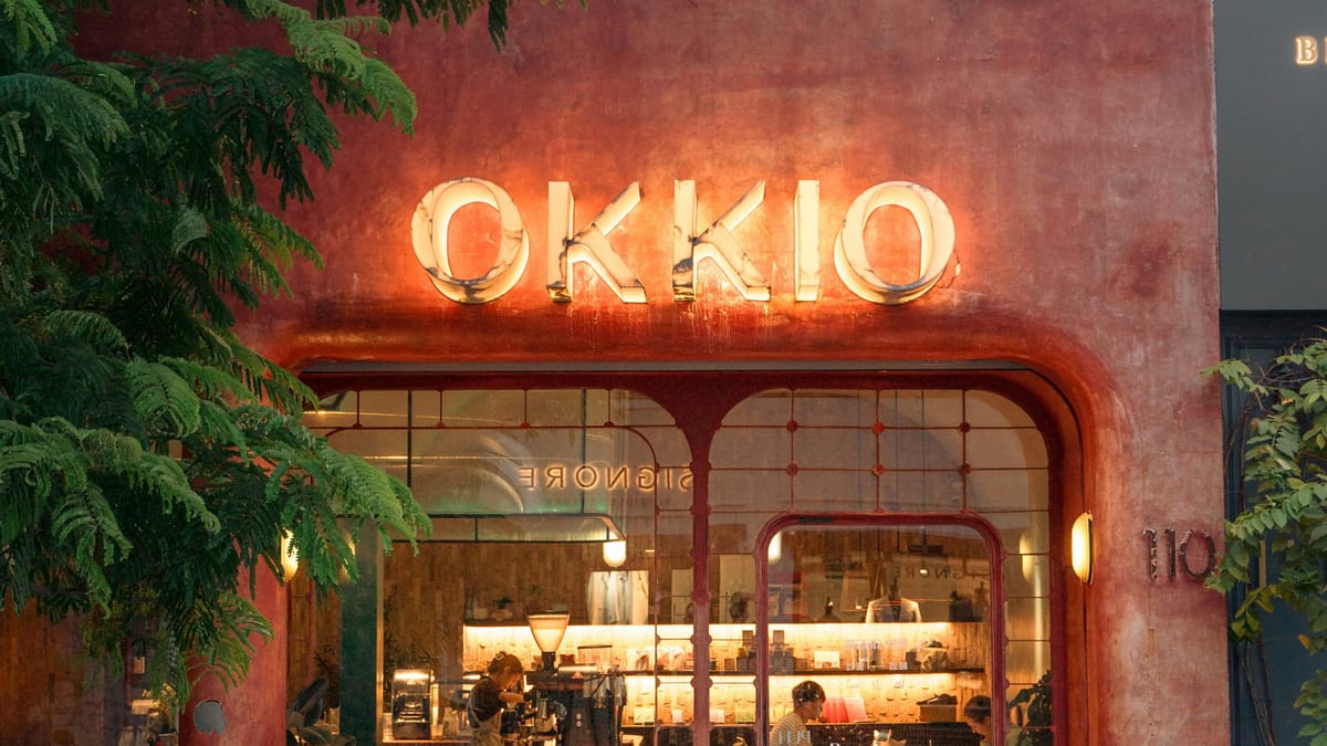 A Vietnamese cafe storefront at night, its OKKIO neon sign glowing warmly, with staff and customers visible through the glass