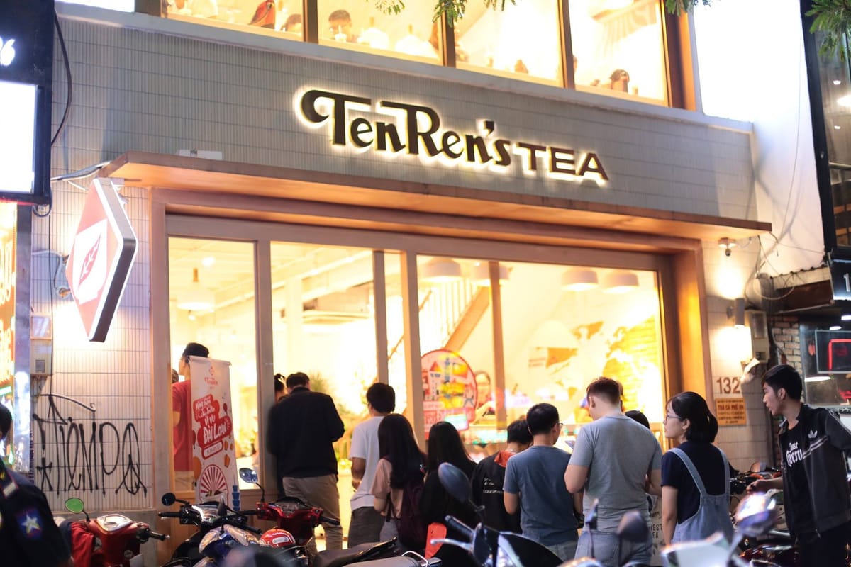 A Ten Ren tea shop in Vietnam at night, the TenRen's TEA sign lit up, with customers gathered outside and motorbikes parked along the street