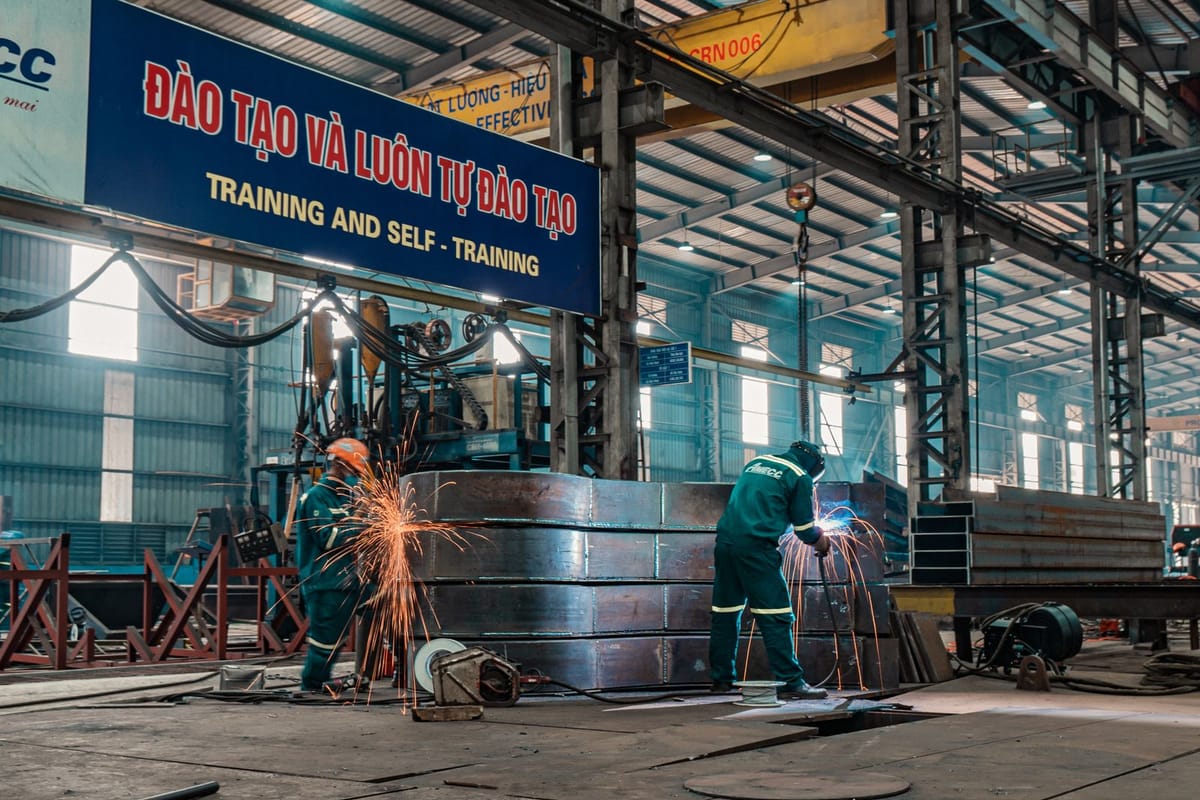 Two workers weld steel components inside a Vietnamese metal fabrication plant, under a Vietnamese-language banner reading "Training and Self-Training"