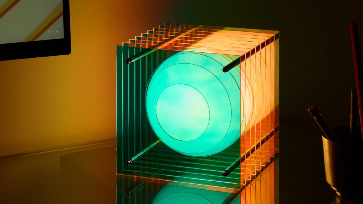 A sculptural table lamp with a milky glass sphere floating between layers of clear acrylic, glowing teal and orange on a dark desk