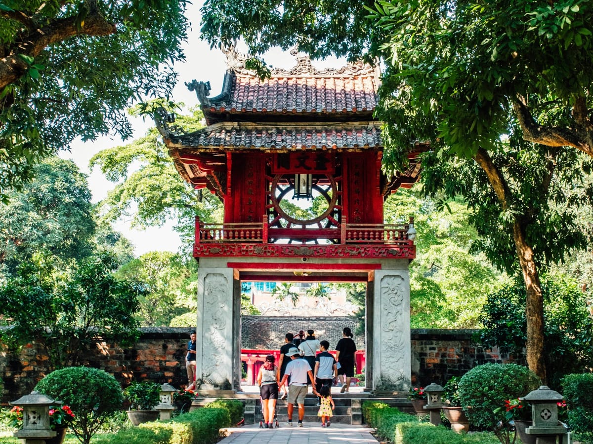 The Khuê Văn Các pavilion at the Temple of Literature in Hanoi, with a family walking along the stone path toward the gate, framed by green trees and red flowers