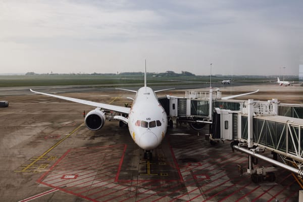 A white widebody jet parked nose-on at a jet bridge, ground crew working under the wing, with aircraft taxiing in the distance