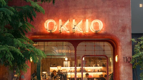 A Vietnamese cafe storefront at night, its OKKIO neon sign glowing warmly, with staff and customers visible through the glass