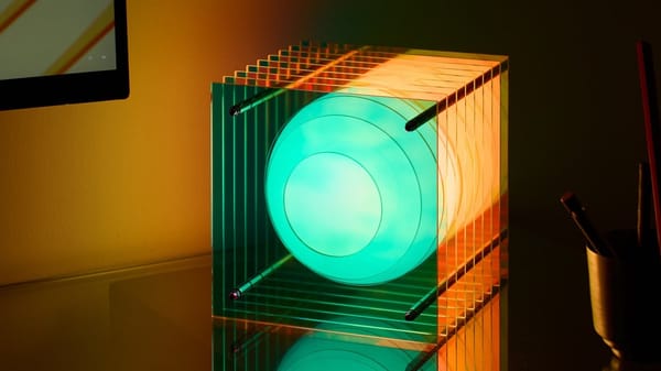 A sculptural table lamp with a milky glass sphere floating between layers of clear acrylic, glowing teal and orange on a dark desk
