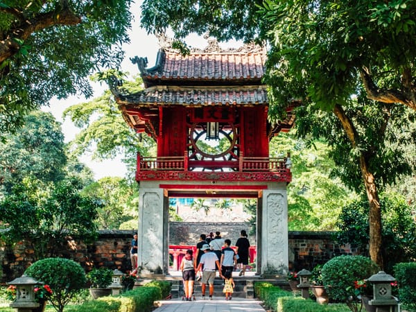 The Khuê Văn Các pavilion at the Temple of Literature in Hanoi, with a family walking along the stone path toward the gate, framed by green trees and red flowers